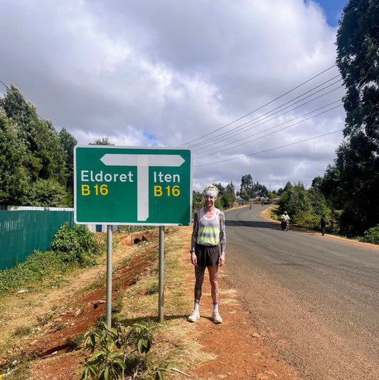 Marathon coach next to Iten road sign training at 2,400m altitude in Iten, Kenya, wearing running gear on a red dirt road.